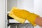 Housewife wearing protective gloves is doing housework. Hand in yellow protective glove is wiping white furniture with rag