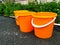 Hree orange plastic buckets of water. Stand in a row on the dark asphalt. On the background of the lawn with green grass and build