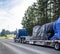 Huge blue classic big rig semi truck transporting covered heavy commercial cargo on step down semi trailer running on the wide