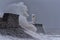 Huge waves breaking against a stone jetty, with a single lighthouse at the end, during a major winter storm.