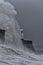 Huge waves breaking against a stone jetty, with a single lighthouse at the end, during a major winter storm.