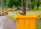 Human hand throw a brown bottle to the yellow color bin on wayside in the garden