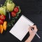 human hand writing spiral notepad with fresh vegetables black wooden backdrop. High quality photo