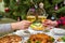 Human hands holding glasses with sparkling wine against beautiful decorated Christmas tree and served table with tasty dishes. New
