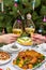 Human hands holding glasses with sparkling wine against beautiful decorated Christmas tree and served table with tasty dishes. New