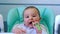 A hungry baby is gnawing on a plastic spoon at the table on a high chair. Teething, whims, itchy gums, introduction of