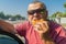 Hungry Caucasian senior driver in sunglasses eating junk food  fried patty near his car while doing short stop on Ukrainian road