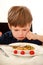 Hungry child sitting in chair at table in kitchen and despises, disapproves of hot meal, because he does not like lunch.