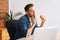 Hungry freelance programmer male eating hamburger watching online video on laptop computer during break, sitting at desk