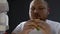 Hungry obese man chewing burger at night near fridge, overeating problem, diet