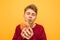 Hungry student in casual clothing is standing on a yellow background and eating fast food, biting a burger with his eyes closed.