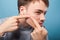 Hygiene and skin problems. Portrait of young brunette man squeezing pimple on face. isolated on blue background