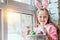 I love Easter!Beautiful little girl in a rabbit costume is sitting at home on the windowsill and holding a basket of Easter eggs