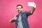 I love selfie! Handsome young man in shirt holding camera and making selfie and smiling while standing against pink background.