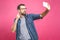 I love selfie! Handsome young man in shirt holding camera and making selfie and smiling while standing against pink background.