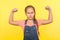 I`m strong! Portrait of brave little girl with braid in denim overalls raising hands showing biceps, feeling power