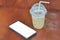 Ice coffee cappuccino, latte, mocha in plastic cup and white straw with water drop near smart phone with white screen for your