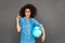 Idea for travel. Mulatto woman standing isolated on grey with terrestrial globe pointing up smiling excited