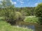 Idyllic landscape of winding river stream meander at lush green meadow with deciduous tree forest, blue sky backgound