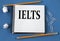 IELTS - word in a white notebook on a blue background with pencils, paper clips