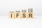 IFSR text written on wooden block with stacked coins on white background