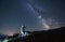 Illuminated man wearing white space suit and helmet sitting on a stool on a hill, pointing at starry sky