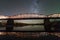 Illuminated metal bridge on concrete supports reflected in water on dark starry sky with Milky Way constellation background. Night