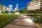 Illuminated pathway concrete sidewalk at park square with downtown Houston skyscraper, capitol tower in background during evening