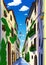 A illustration of a narrow village street with warm-colored houses, greenery, and a blue sky
