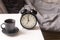 Image alarm clock with black cup of coffee on a white bedside table in front of the bed with gray linens. The room is in beige