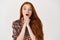 Image of amazed redhead woman gasping with amazement and interest, looking at camera, standing over white background