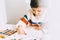 Image of artistic little girl painting with oil pencils, sitting at white desk at home. Pretty smiling preschool kid drawing