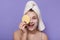 Image of beautiful smiling woman covers her eye with sponge, wearing white towel on head, stands against lilac background, keeps