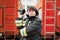 Image of brunette firefighter with camera at hand standing next to firefighters car.