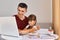 Image of brunette young adult father sitting with schoolgirl daughter at table and looking at laptop monitor, watching online