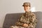 Image of Caucasian smiling soldier woman wearing camouflage uniform sitting on sofa and looking away with positive happy facial