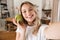 Image closeup of joyful blond woman laughing and holding green apple while taking selfie photo in living room