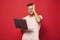 Image of confused young man in striped t-shirt smiling and holding gray laptop, isolated over red background