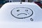 Image of a crying face emoji on the snow on the back window of a car on a bright winter day