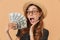 Image of excited european woman 20s wearing straw hat and sunglasses screaming and demonstrating fan of dollar banknotes, isolate