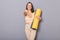 Image of friendly sporty woman wearing beige tracksuit holding yoga mat  on gray background, pointing to you, selecting