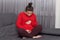 Image of future mother has hair knot, keeps legs crossed, wears red jumper and leggings, keeps both hands on belly, sits on sofa,