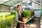 Image of gardener 20s wearing apron standing with plants in hands while working in greenhouse