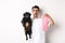 Image of hipster guy pet owner, holding cute black pug and dog poop bag, standing over white background