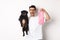 Image of hipster guy pet owner, holding cute black pug and dog poop bag, standing over white background