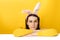 Image of lovely female leans on white table, wears fluffy ears, dressed in sweater, models over yellow background