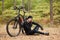 Image of male mountain biker resting on cycling trip, sitting on ground under tree with his bicycle lying next to him and his