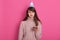 Image of offended upset young woman wearing pink sweater and birthday hat, holding cake with candle with unhappy facial expression