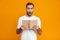 Image of puzzled man 30s in white t-shirt holding and reading book,  over yellow background