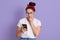 Image of scared brunette female with knot and headband biting her finger, feels fear, holding smart phone, wears white t shirt,
